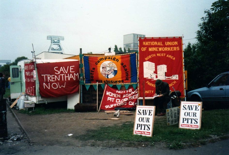 Colour photograph of a white caravan with a green horizontal stripe, covered in red banners with white text reading “Save Trentham Now” and “North Staffs Miners’ Wives Action Group”. The caravan is parked on grass and dirt,  surrounded by several other banners standing nearby.